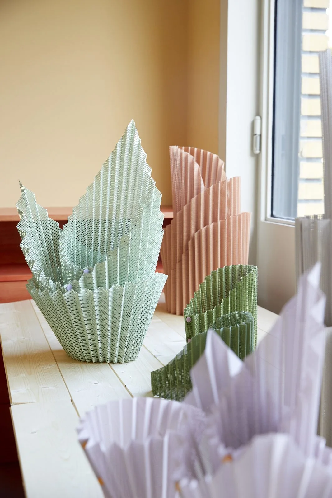 Structures en papier plié, vert d'eau et rose, du projet DÉPLI, posées devant une fenêtre.