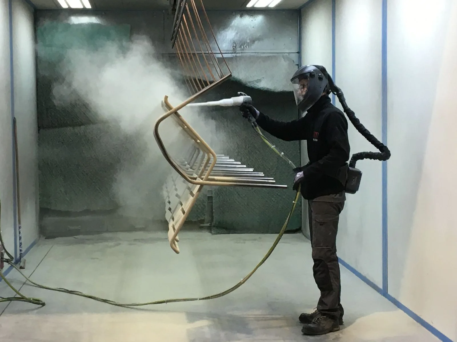 Opérateur en cabine de poudrage appliquant la finition sur un piètement de chaise, dans l'atelier de rm-mobilier.