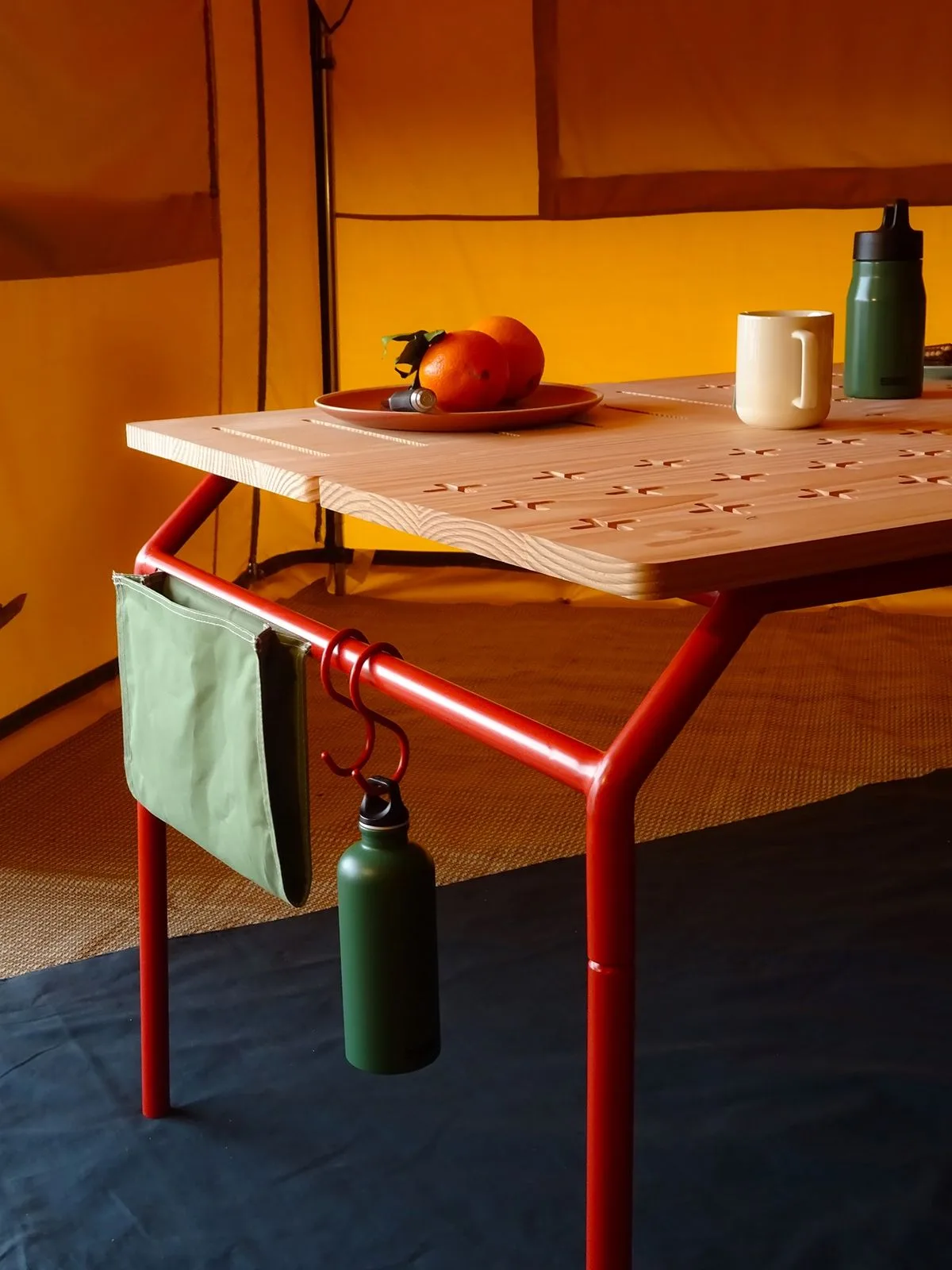 Photo officielle de la gamme : table à piètement rouge et plateau de bois ajouré, sacoche suspendue.
