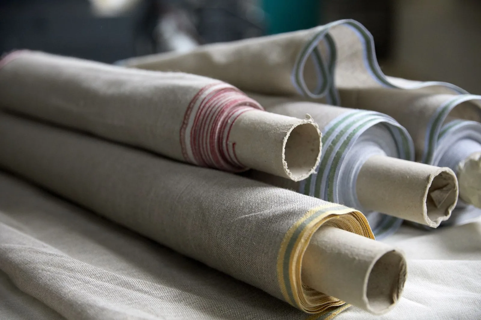 Rouleaux de toile de lin enroulés côte à côte, liserés bleus, rouges et jaunes visibles en bout.